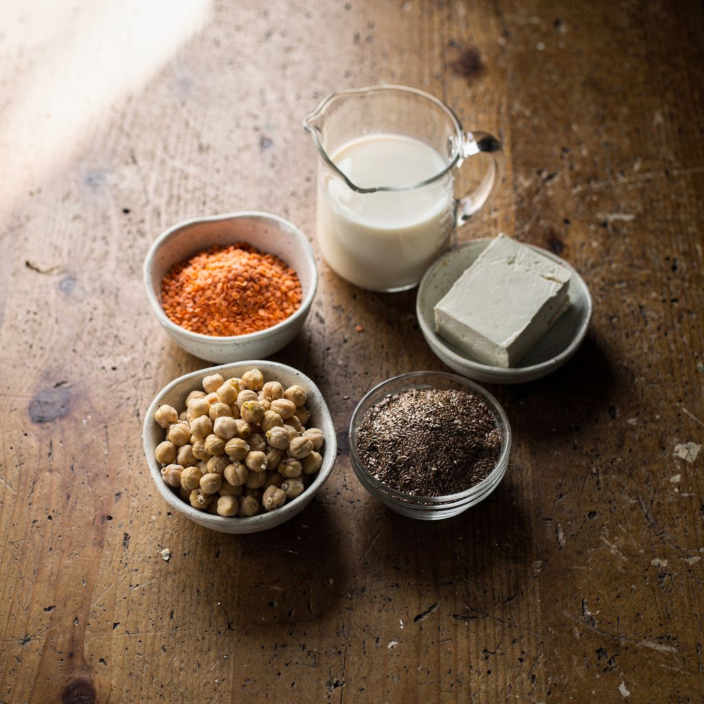 A wooden kitchen table seen from above with small bowls of red lentils, chickpeas, a block of tofu, flax seeds and a glass jug of soy milk arranged in a loose circle, with calm empty space around them.