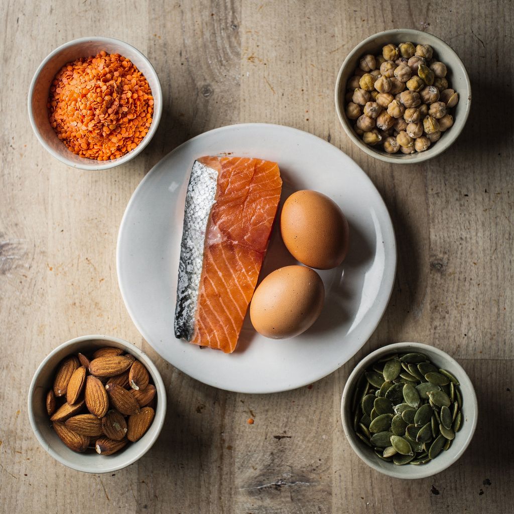 An overhead arrangement of small bowls holding dried red lentils, chickpeas, almonds and pumpkin seeds, set around a plate with a raw salmon fillet and two brown eggs, on a plain wooden kitchen table with empty space around the edges.