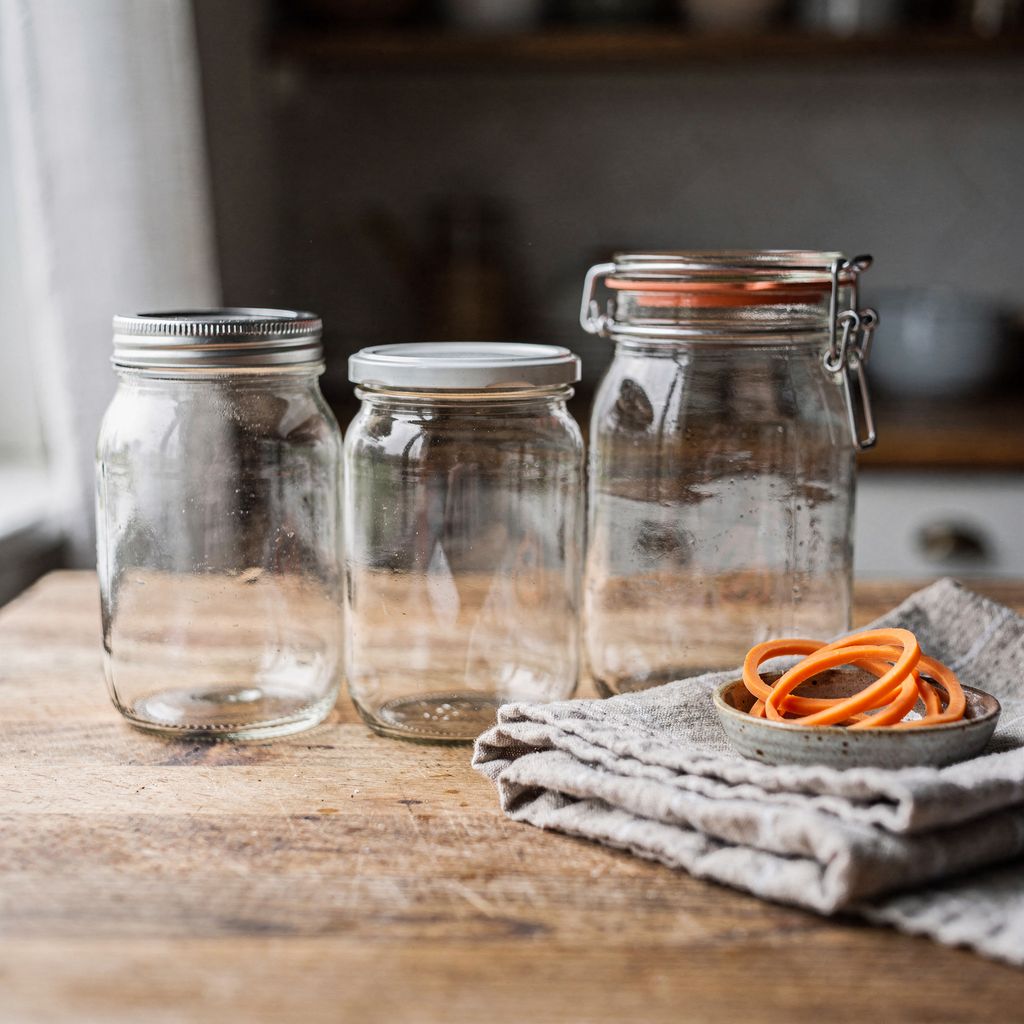 Three empty glass preserving jars standing side by side on a wooden kitchen counter — one with a screw-on metal lid, one with a flat press-on lid, and one with a glass lid held by a wire clamp and a rubber ring — with a folded cloth and a small bowl of spare rubber rings beside them and calm empty s
