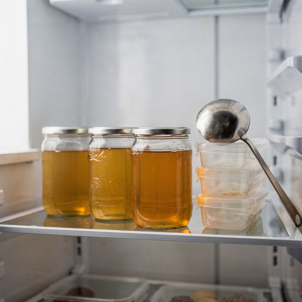 Three lidded glass jars of clear golden broth standing on a clean refrigerator shelf, with a small stack of empty storage containers and a ladle resting beside them.