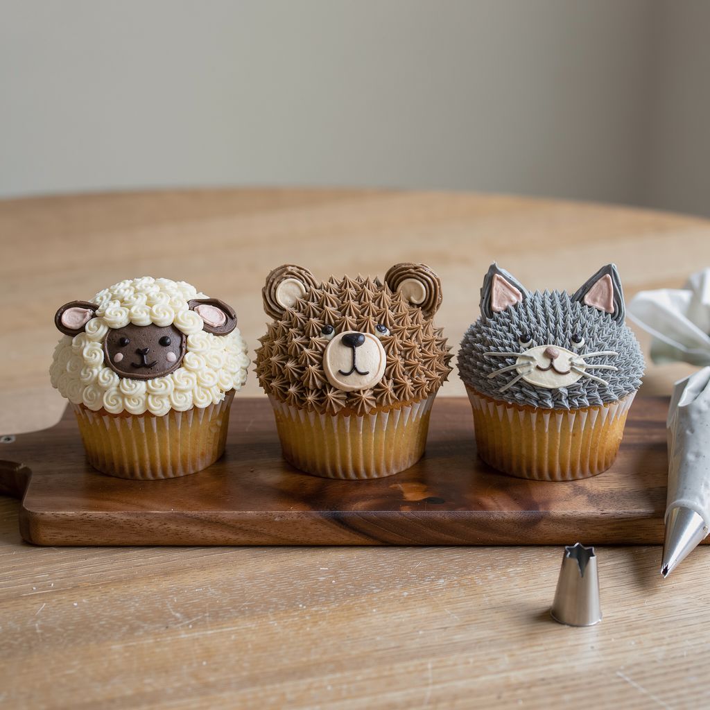 Three frosted cupcakes in a row on a wooden board: one topped with curly white buttercream sheep wool and a dark face, one with brown star-piped bear fur and round ears, one with a grey buttercream cat face with pointed ears and fine whiskers. A piping bag lies beside them with calm empty space arou