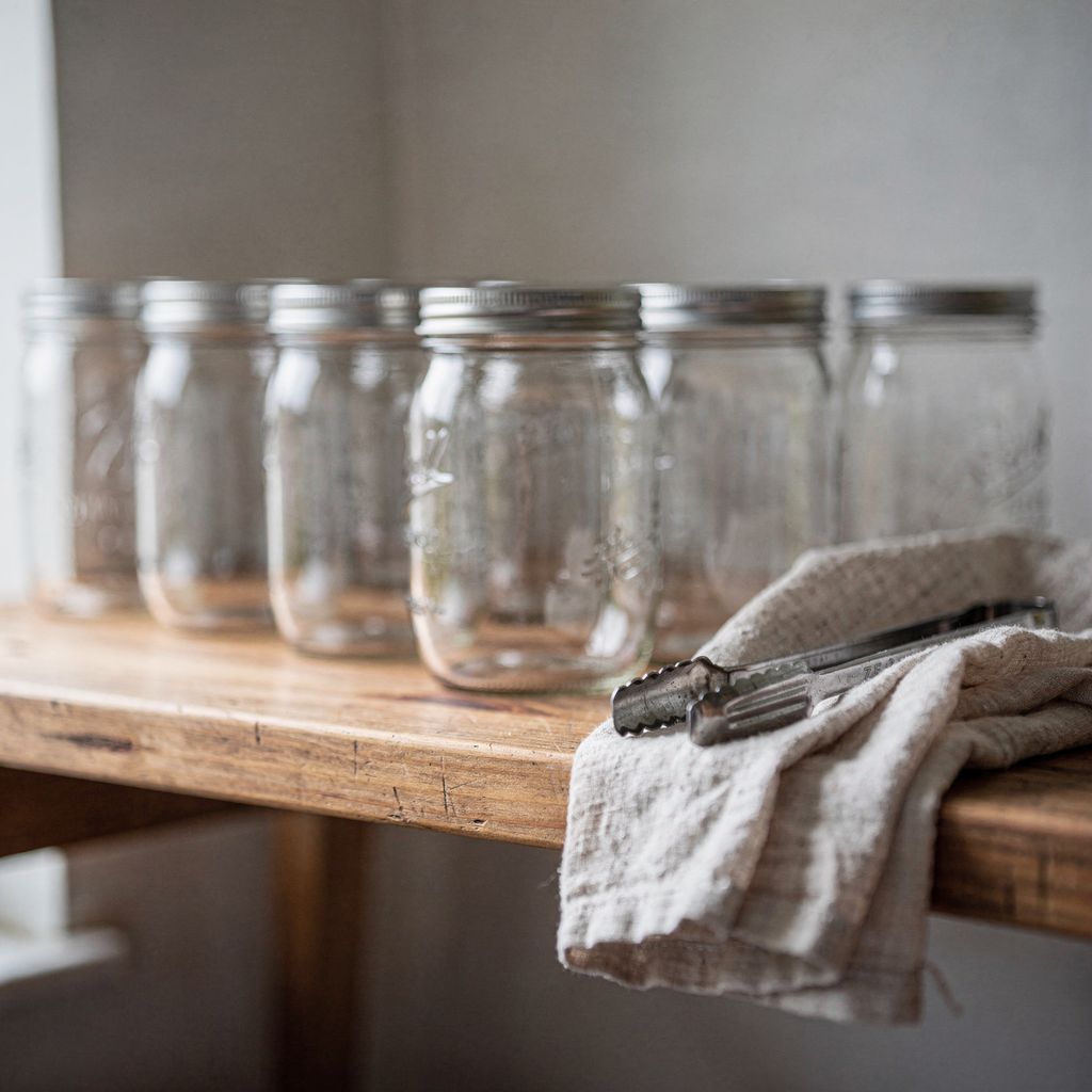 A wooden pantry shelf lined with rows of closed glass preserving jars with plain metal lids, a folded linen cloth and a pair of jar tongs resting beside them, centred in a square frame with calm empty space around the shelf.