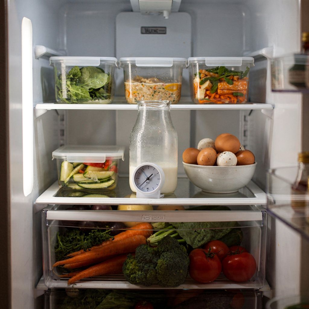An open refrigerator seen straight on, shelves neatly filled with glass storage containers, a jug of milk, a bowl of eggs and a crisper drawer of vegetables, with a small round fridge thermometer resting on the middle shelf.