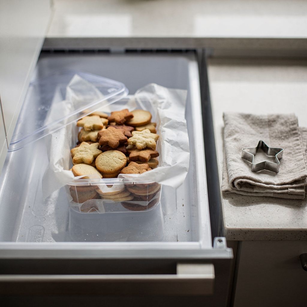An open freezer drawer holding a stacked clear container of small round and star-shaped Christmas cookies separated by folded parchment layers, with a folded kitchen towel and a metal cookie cutter resting on the counter beside it, centred in a square frame with calm space around.