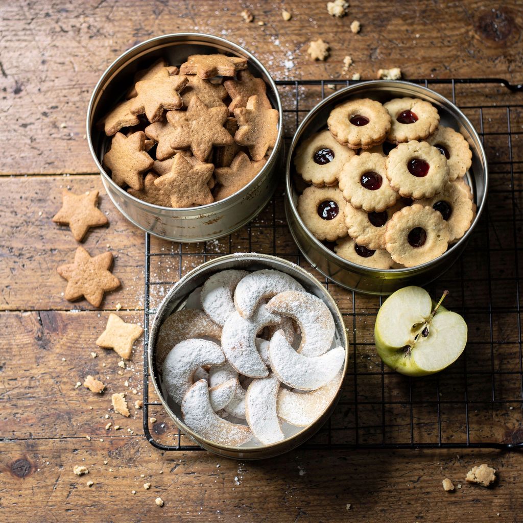 An overhead view of a wooden kitchen table with three open metal tins of assorted Czech Christmas cookies at different stages — plain baked gingerbread shapes, jam-sandwiched linzer rounds, and sugar-dusted crescents — beside a halved apple and a cooling rack. Subject centred in a square frame with