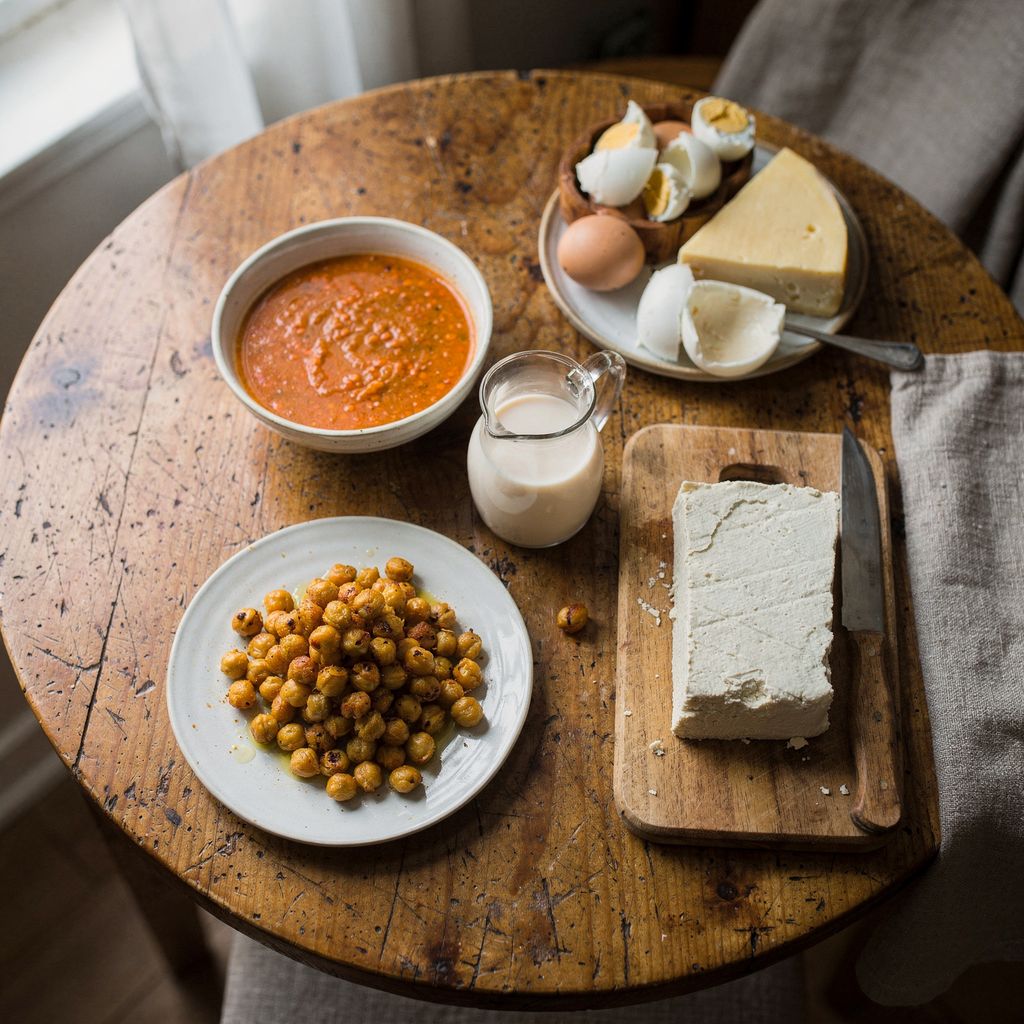 A round wooden table seen from above with a spread of dishes: a bowl of red lentil soup, a plate of roasted chickpeas, a jug of almond milk, a block of tofu on a small board, and slightly apart a dish of boiled eggs and a wedge of cheese. Centred in a square frame with calm empty space around the ed